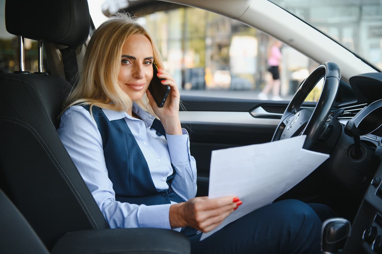 Mulher sentada no banco de motorista do carro, segurando o volante com a mão esquerda e um punhado de papéis com a mão direita. Ela olha para a frente com um sorriso discreto.