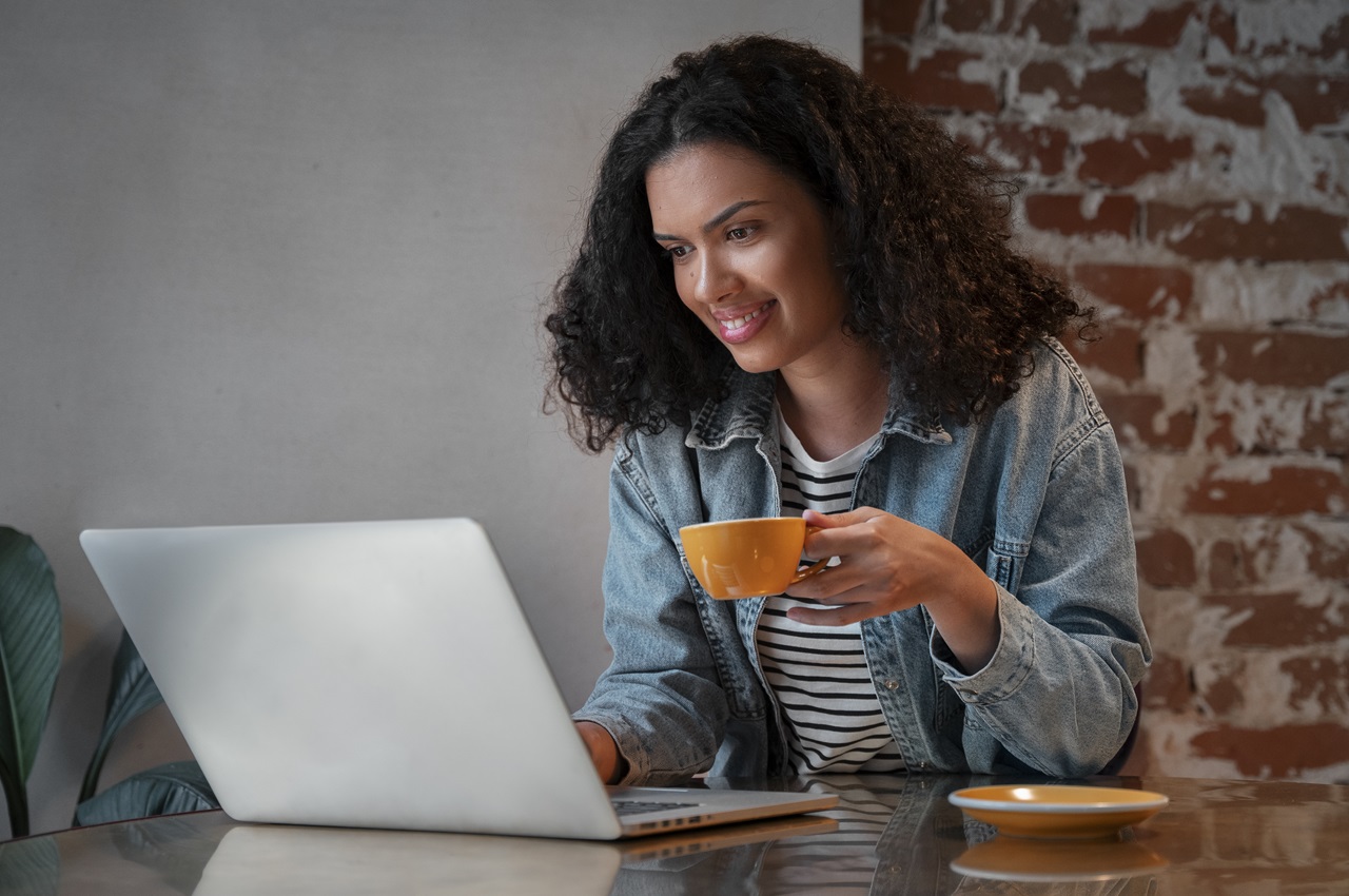 Mulher de cabelos castanhos encaracolados, sentada na mesa do escritório, utilizando um computador e segurando uma xícara de café amarela.