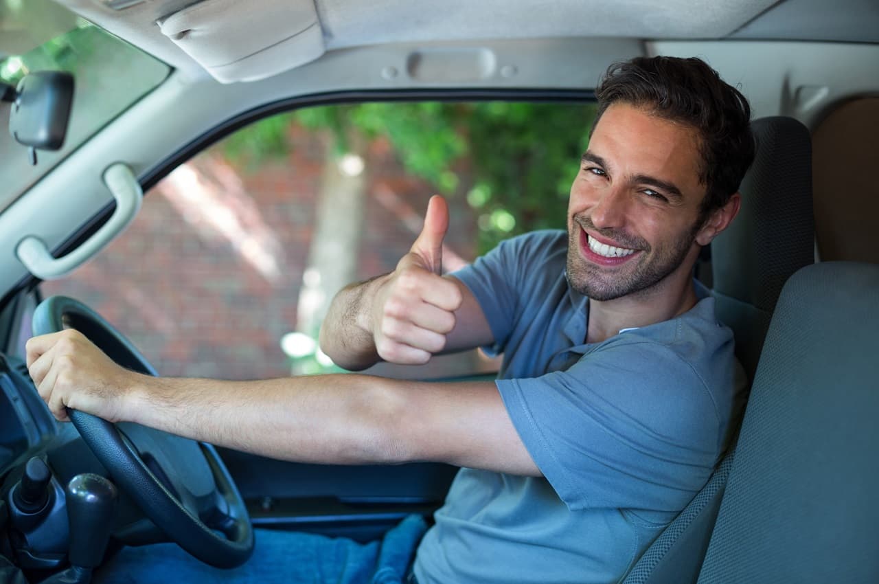 Homem sorridente, vestindo camiseta cinza simples, dentro de um carro e sentado no banco de motorista. Ele faz joia com a mão esquerda e segura o volante com a mão direita.