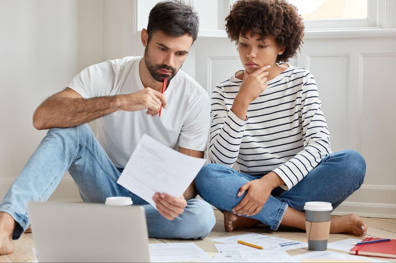 Homem e mulher com expressões concentradas, sentados no chão de um quarto e olhando para um papel com contas a pagar.