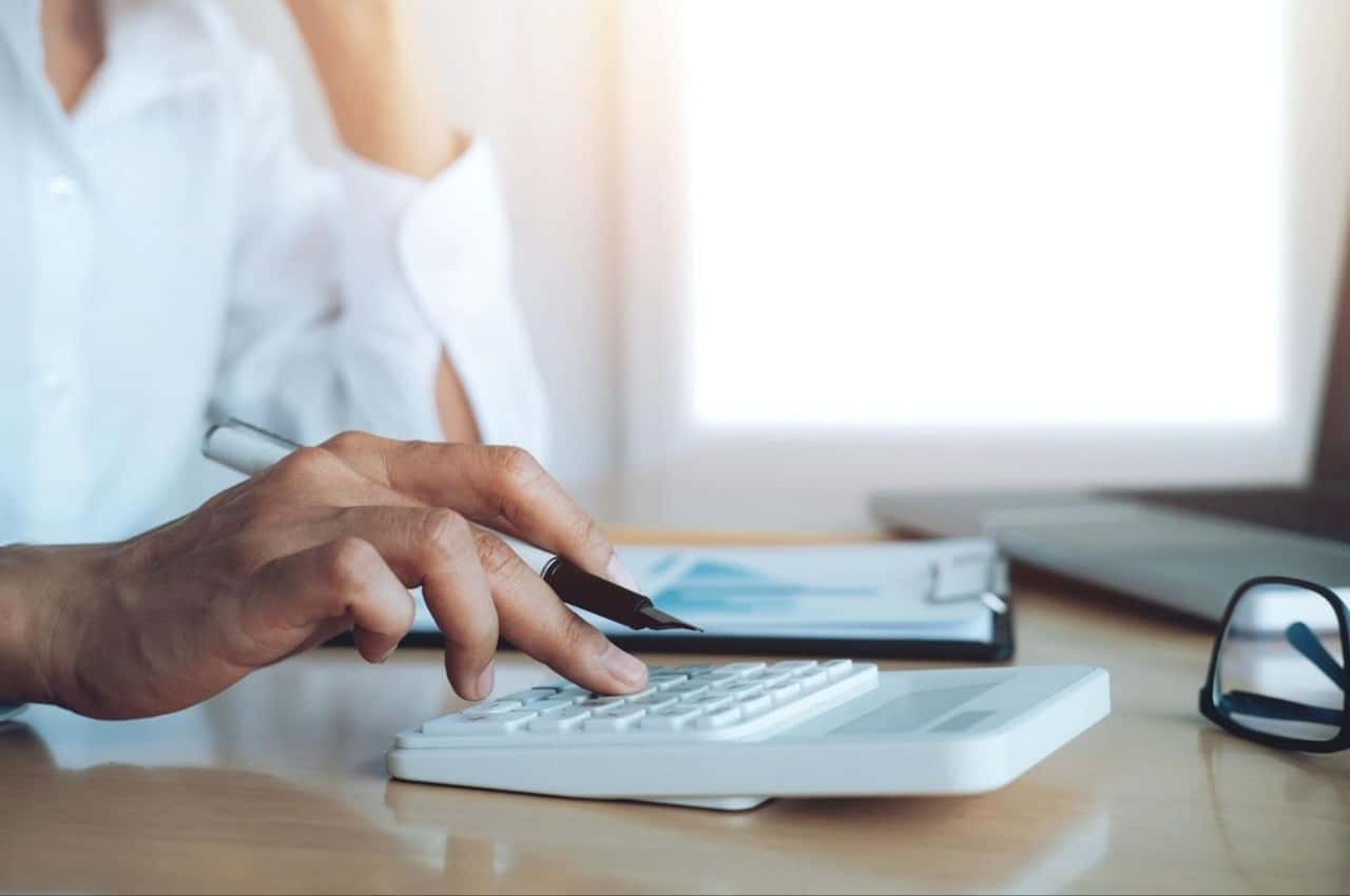 Pessoa digitando em uma calculadora e segurando uma caneta, com a mão esquerda ela apoia o queixo.