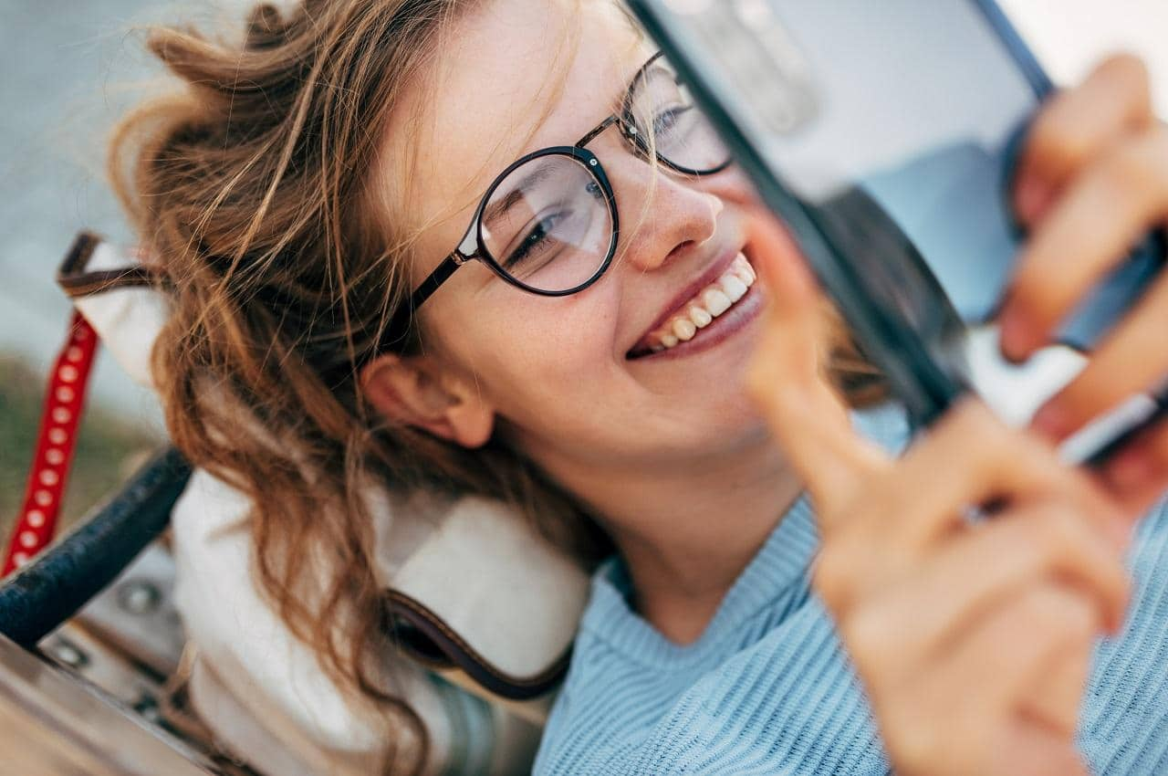 Mulher sorridente com óculos de grau, deitada de barriga para cima e segurando o celular acima do rosto.