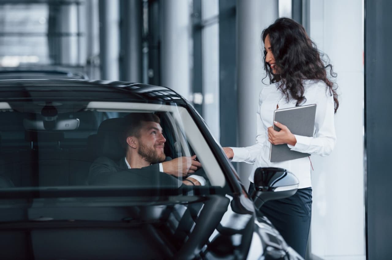 Homem de blazer sentado no banco do motorista de um carro preto, pegando o objeto da mão de uma mulher que está de pé ao lado do carro. A mulher está vestindo uma camisa branca e calça preta, segurando uma pasta cinza com uma mão, e entregando um objeto para o homem pela janela do carro.