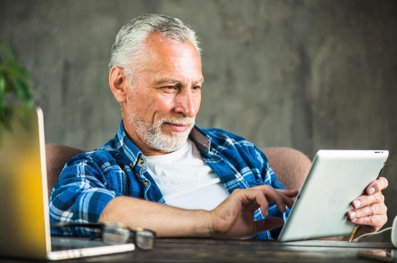 Homem sênior com cabelos grisalhos e utilizando um tablet em seu escritório. Ele veste camisa branca básica e blusa xadrez azul.