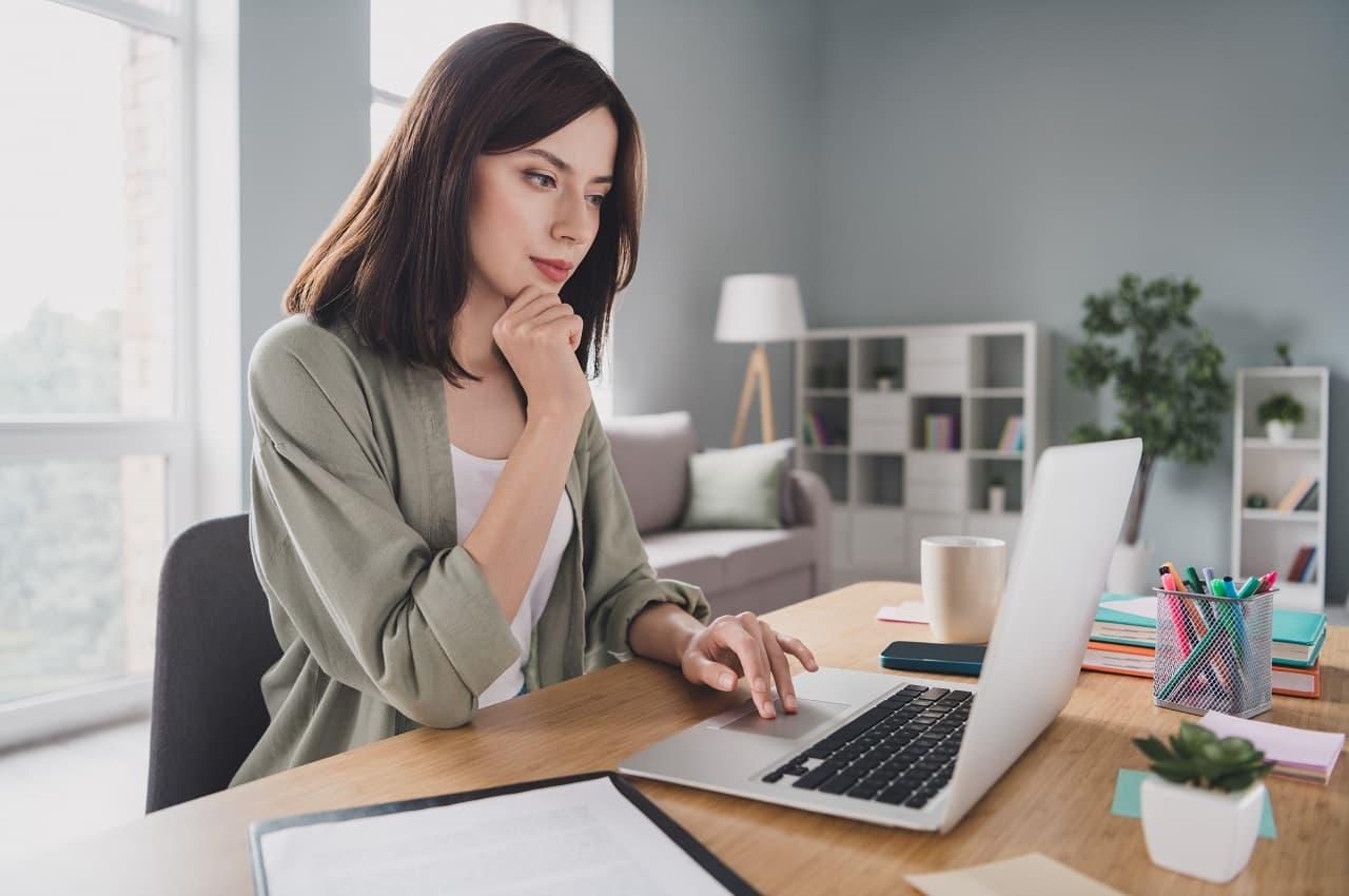 Mulher de cabelos curtos castanhos digitando no computador no escritório. Ela veste uma blusa verde e uma camisa branca básica.