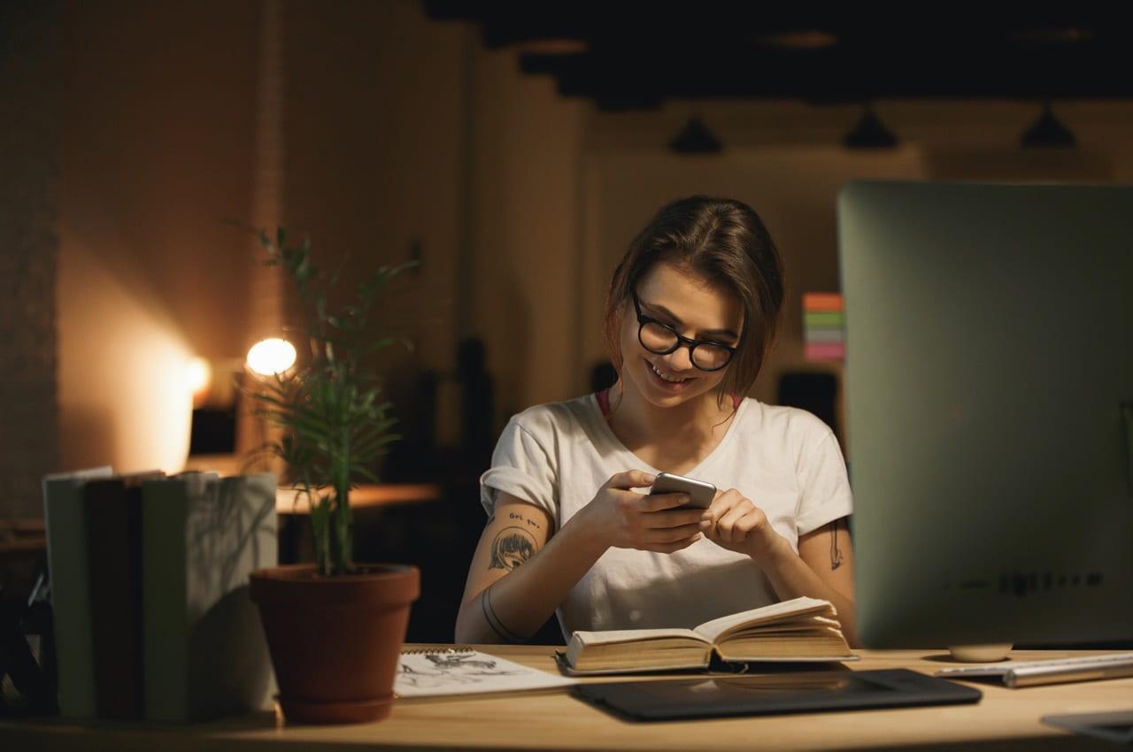 Mulher sorrindo enquanto utiliza o celular em um ambiente de trabalho noturna.