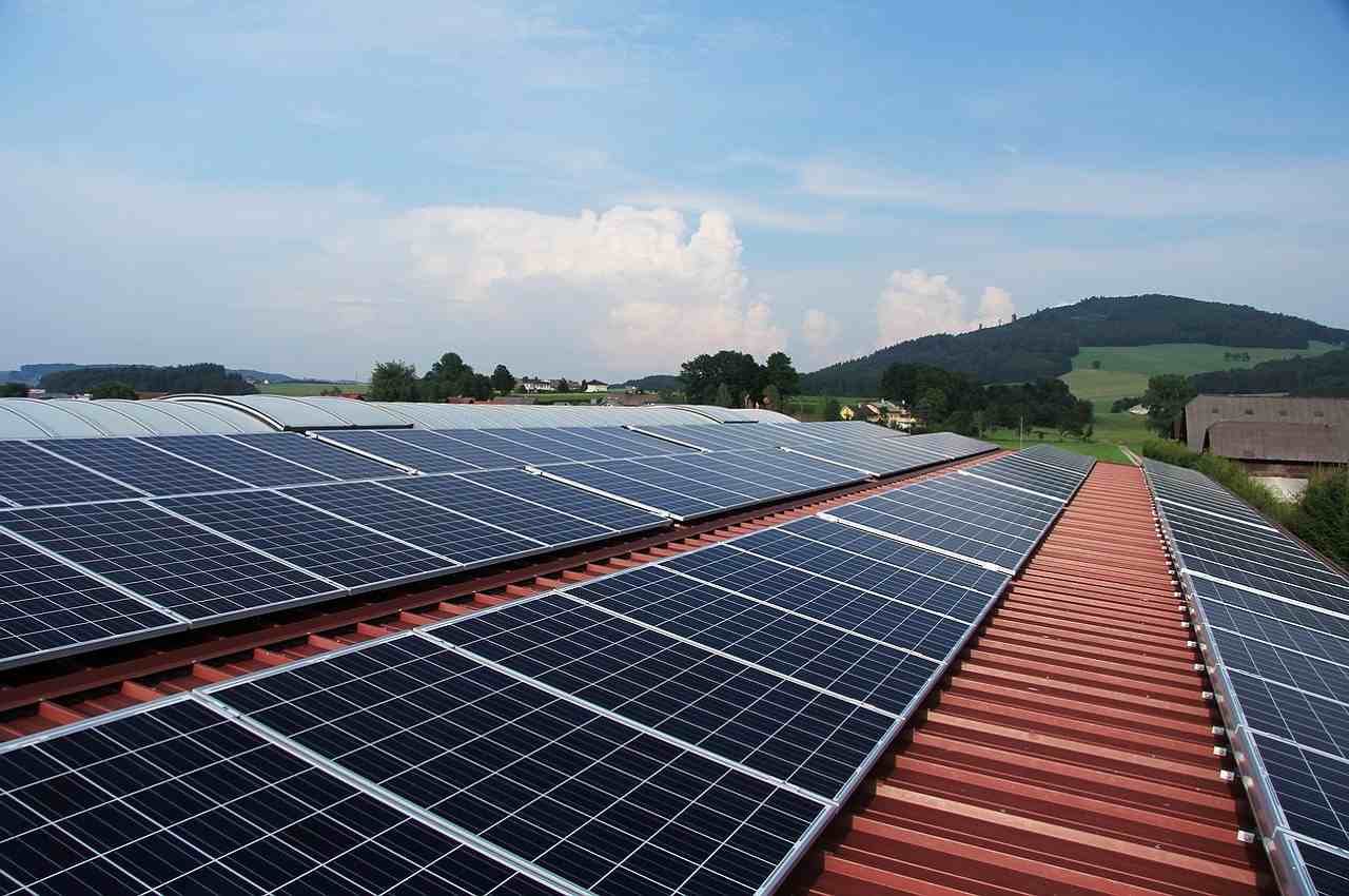 Telhado de galpão grande com várias placas solares instaladas. Ao fundo existe outros galpões, mas sem energia solar instalada. 