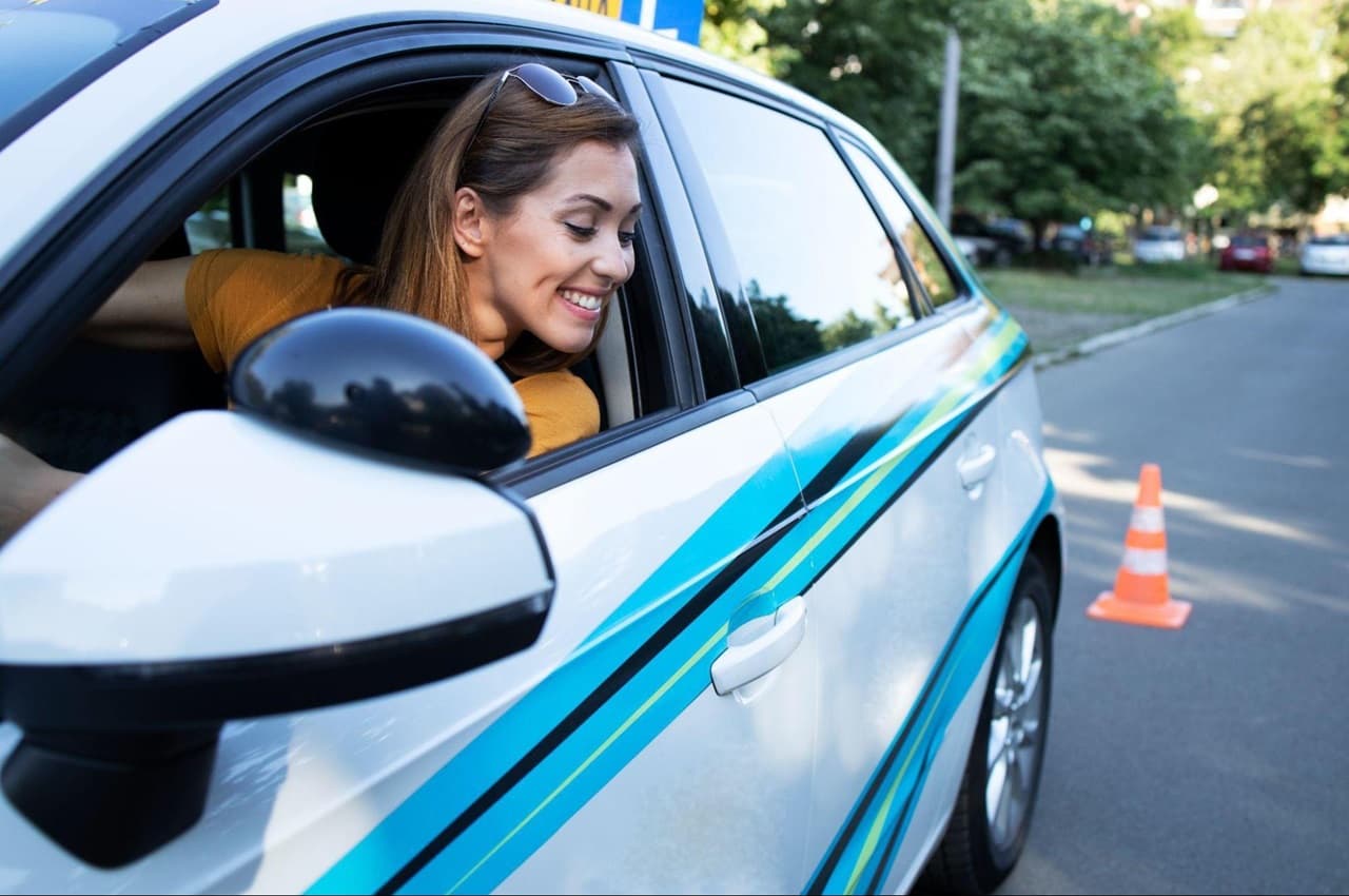 Mulher sorridente renovando CNH em prova prática de veículo. Ela olha pela janela de um carro azul próximo aos cones de sinalização.