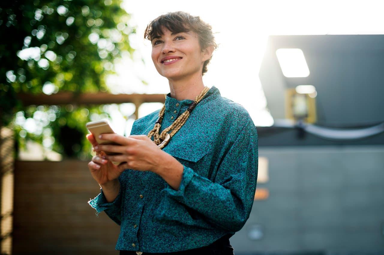 Mulher sorridente segurando um celular com as duas mãos, enquanto reflete sobre a averbação de empréstimo consignado.