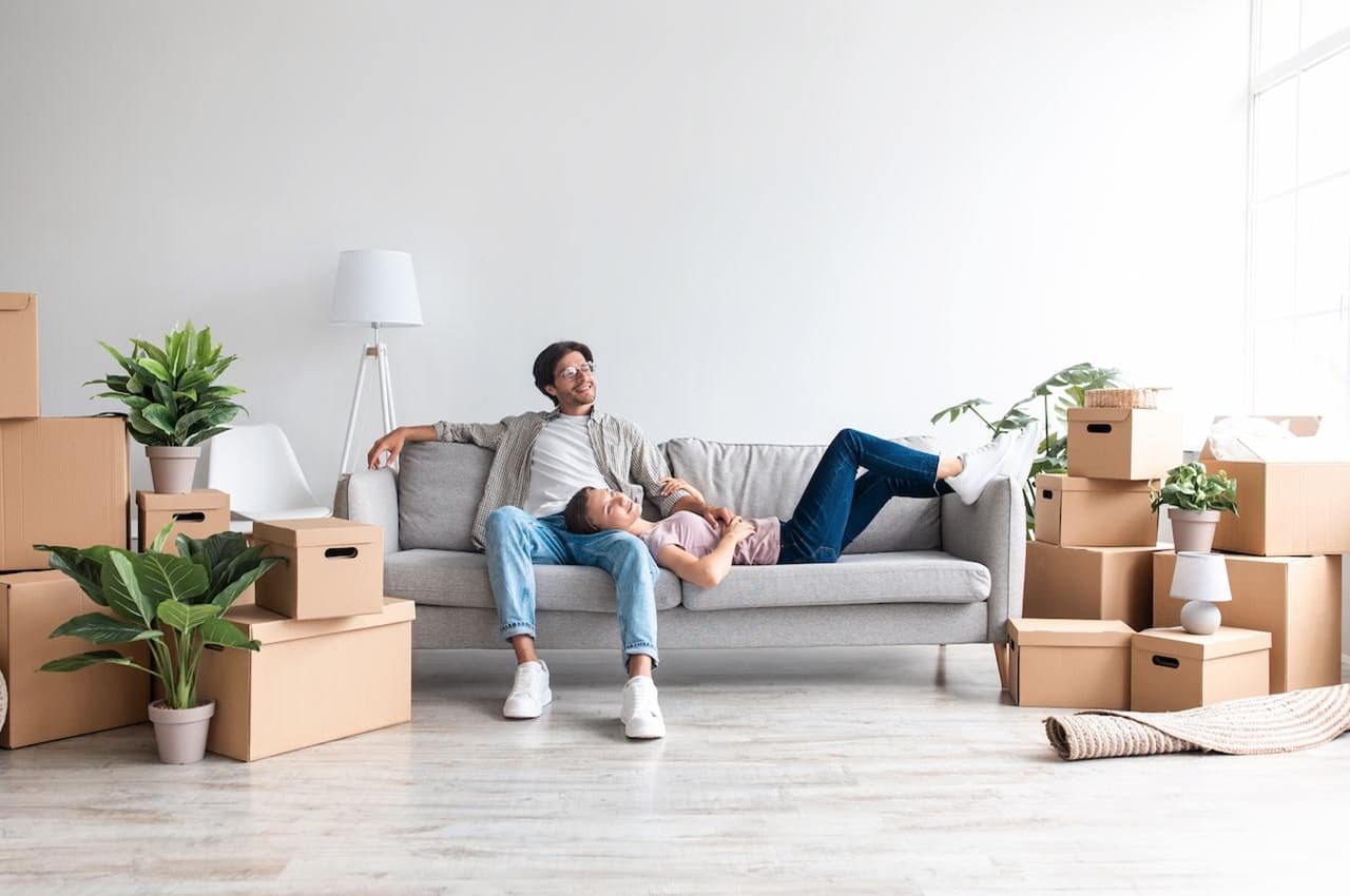 Casal sorridente de noivos (homem e mulher) sentados no sofá de sua casa nova e desempacotando seus pertences que estão em caixas.