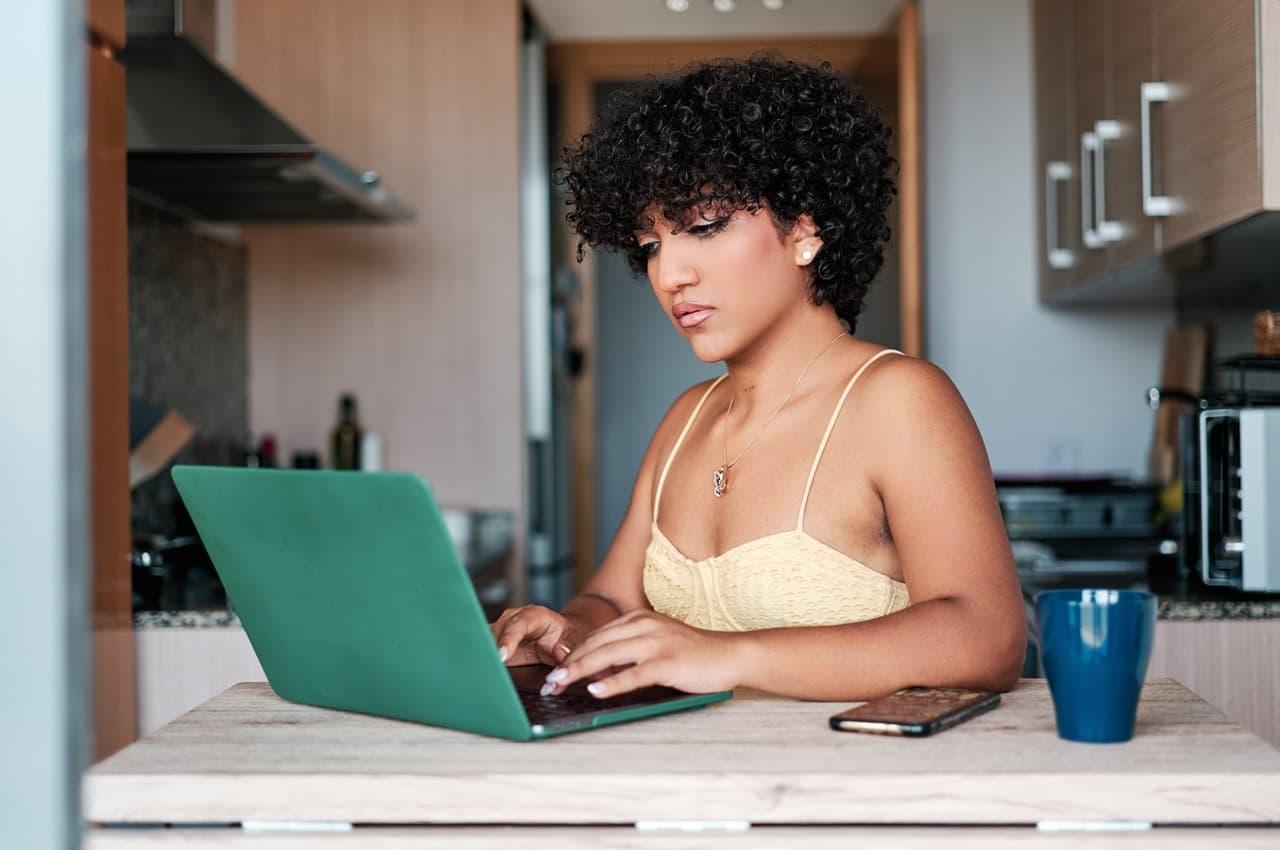 Mulher com expressão concentrada, usa laptop verde para acessar financiamento e empréstimo ao mesmo tempo. A mulher veste regata amarela.