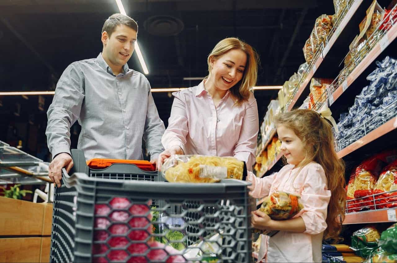 Família sorridente de mulher, homem e criança pequena no supermercado realizando compras do mês com o carrinho cheio de mercadorias.