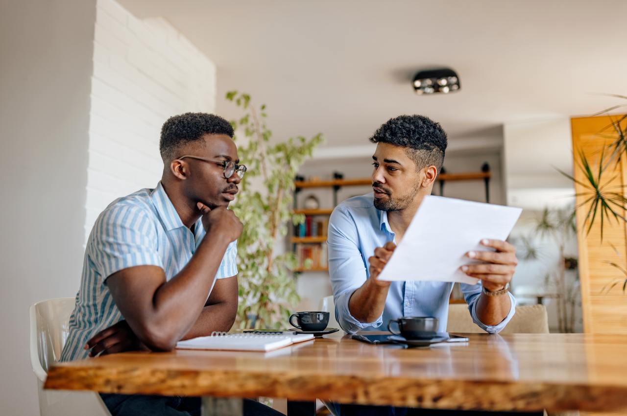 A imagem mostra dois homens sentados, tomando café e analisando alguns papéis.