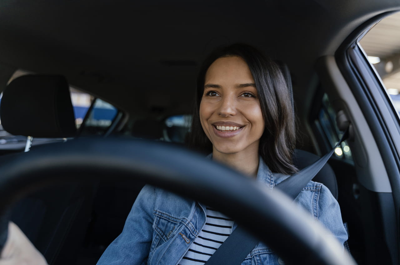 Mulher sorridente de cabelos castanhos curtos dirige veículo e veste camiseta jeans e blusa listrada.