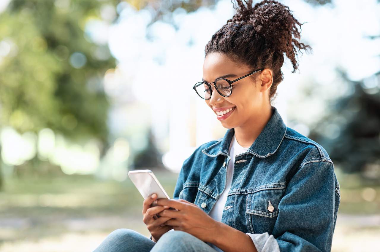 Mulher negra sorridente, de cabelos presos e blusa jeans, digitando no celular sobre CDI em um parque.