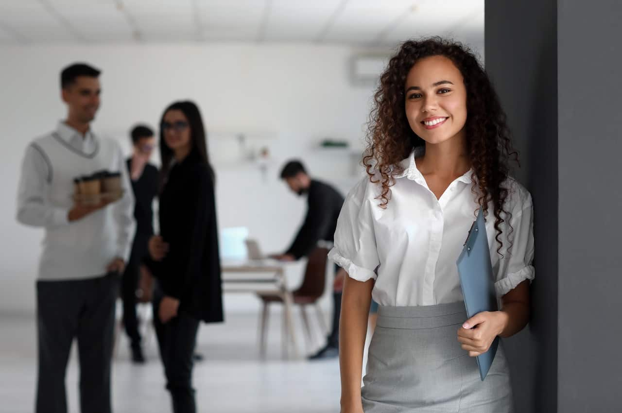 Mulher sorridente de cabelos cacheados longos, vestindo blusa e saia social. A mulher segura uma pasta de documentos e atrás dela tem um grupo de colegas de trabalho.