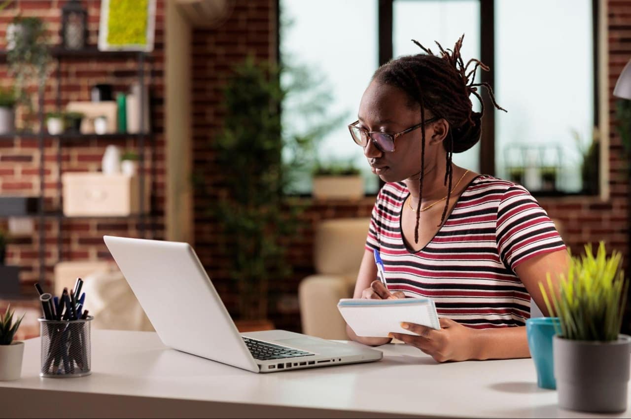 Mulher com expressão concentrada segura papel nas mãos e acessa um laptop em sua casa. A mulher usa o cabelo em coque e veste camisa listrada vermelha e branca.