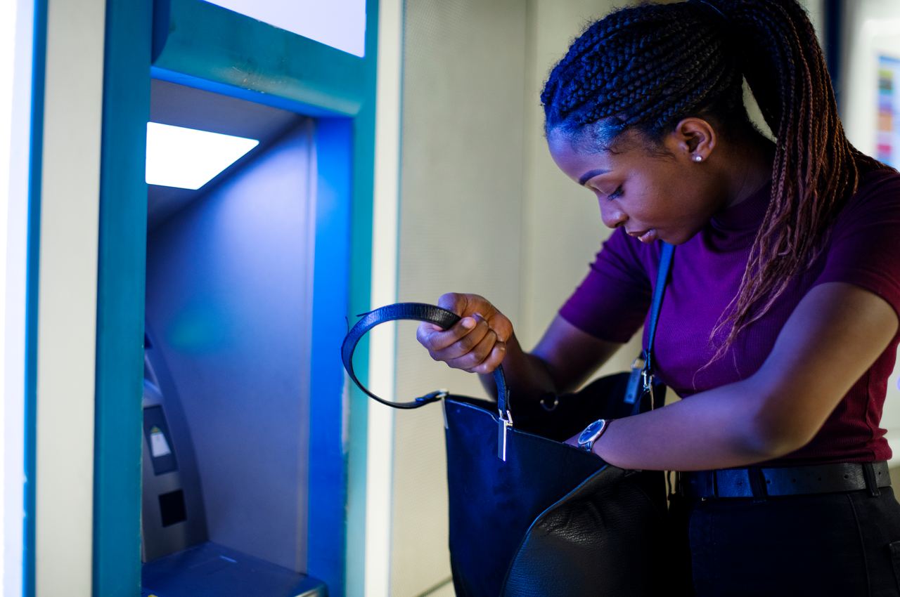 A imagem mostra uma mulher em um caixa eletrônico. Ela está procurando algo em sua bolsa.