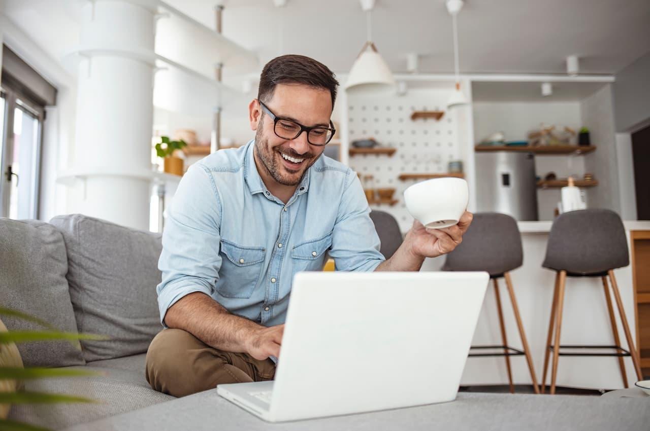 Homem sorridente veste camisa jeans, óculos de grau e pesquisa saldo parcial FGTS pelo laptop que está em cima de uma mesa em frente ao sofá.