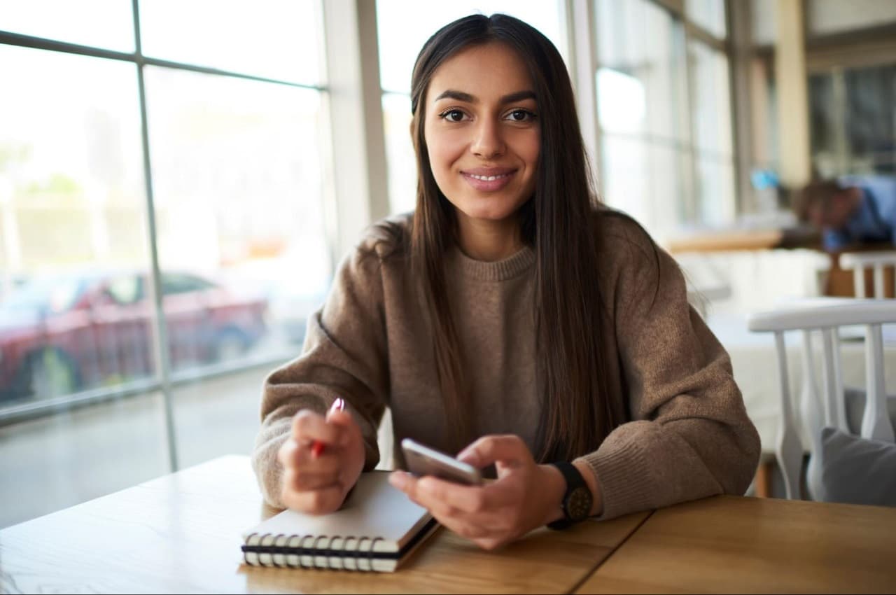 Mulher sorridente de cabelo castanho na cafeteria, utilizando celular para antecipar saque-aniversário e realizando anotações em bloco de notas branco.