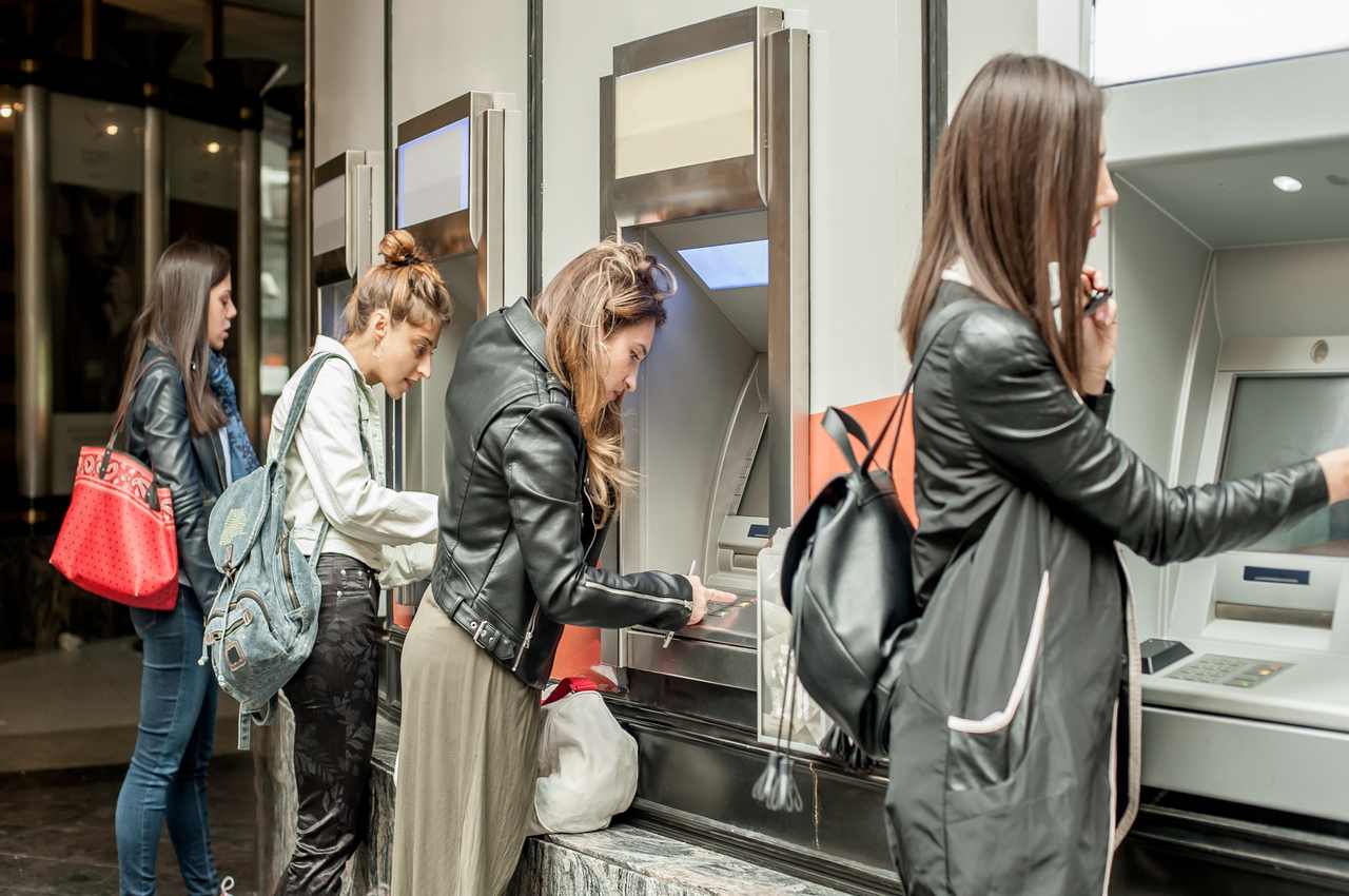 A imagem mostra quatro mulheres em um banco. Cada uma está usando um caixa eletrônico.