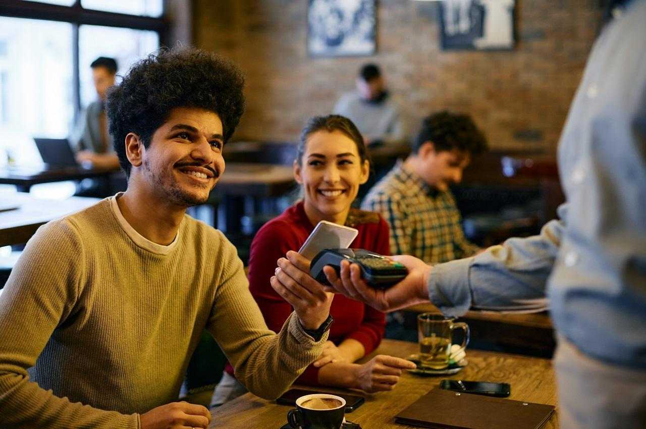 Homem sorridente com suéter mostarda, pagando com aproximação uma conta de um restaurante. Ao seu lado, tem uma mulher sorrindo para o homem.