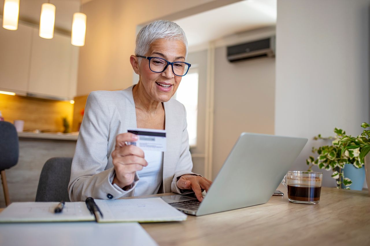 A imagem mostra uma mulher fazendo compras em seu laptop, com seu cartão de crédito.