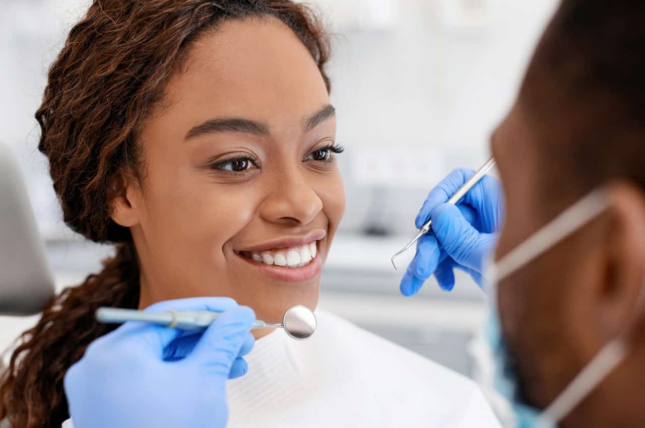 Mulher sorridente realizando uma limpeza dental com dentista paramentado (a pessoa não aparece na imagem).