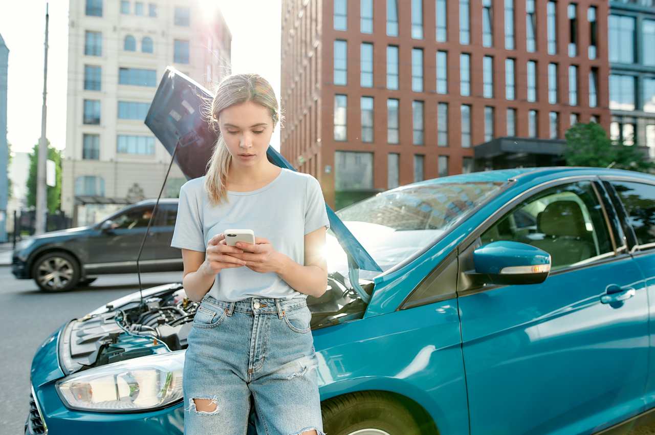 Mulher loira com expressão preocupada, utilizando celular para acionar seguro auto. Atrás da pessoa, tem um carro com capô frontal aberto.