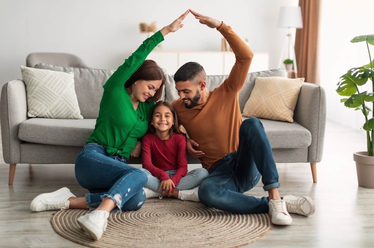 Família sorridente sentada no tapete da sala, composta por uma mulher de blusa verde, um homem de blusa marrom e uma criança de blusa vermelha. O casal faz uma casa com as mãos. 