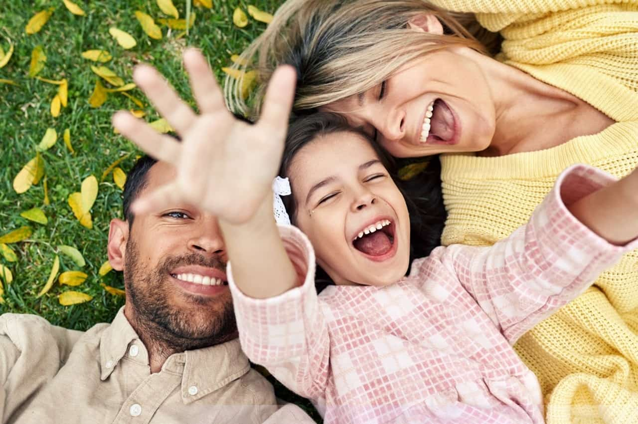 Família sorridente de homem, mulher e uma criança com os braços para cima e deitados no gramado de uma praça arborizada.