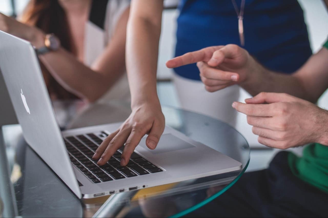 Grupo de pessoas ao redor de uma mesa e utilizando um laptop. Os rostos das pessoas não aparecem na imagem.