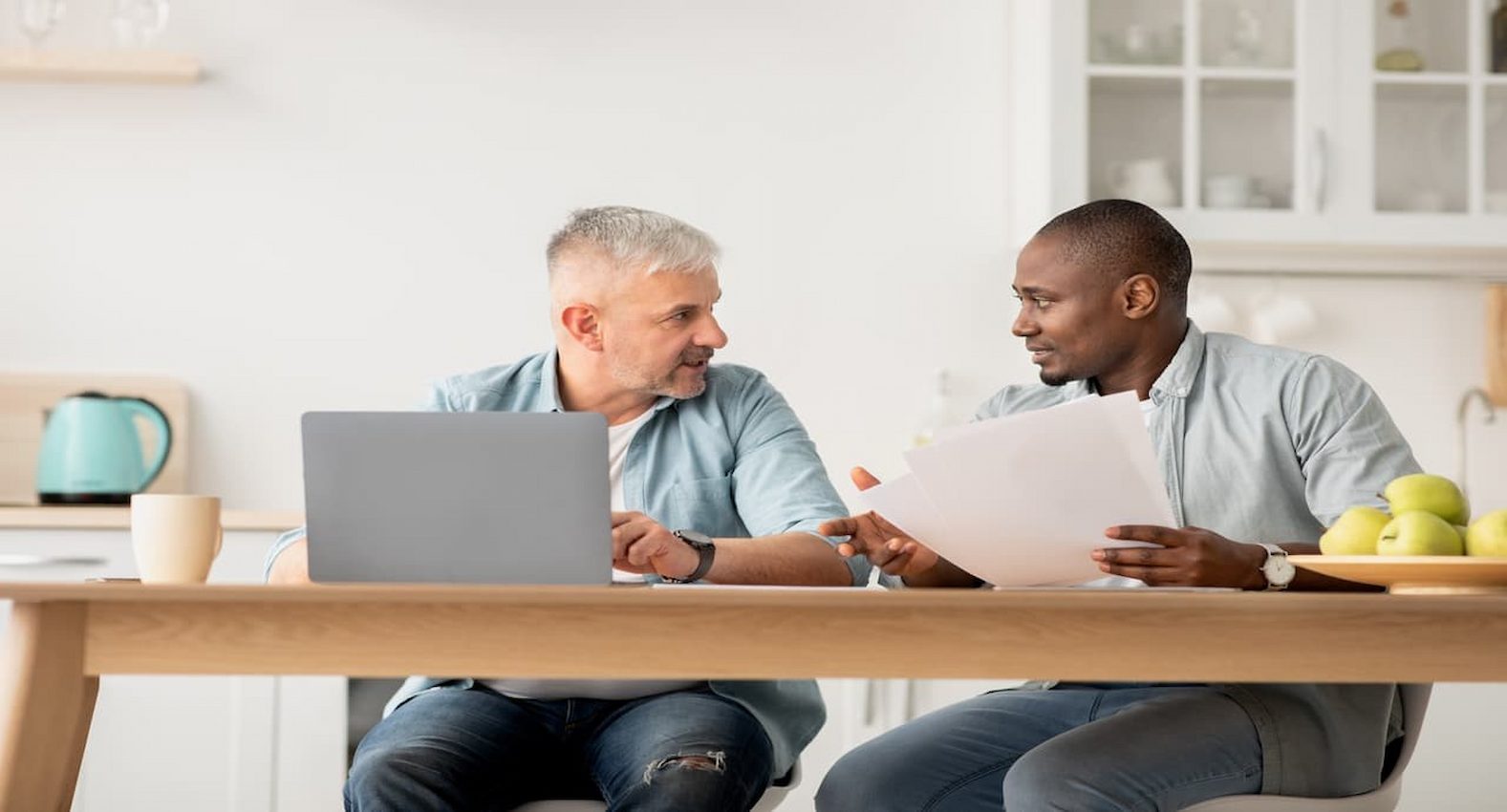 A imagem mostra dois homens sentados à uma mesa, um está usando um laptop e outro segurando alguns papéis.