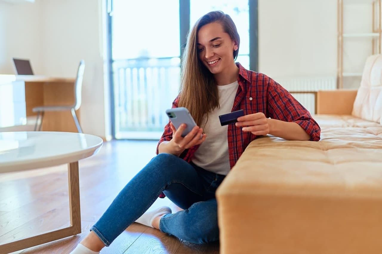 Mulher sentada no chão de madeira da sala de estar, vestindo meias, calça jeans e blusa xadrez vermelha. Ela utiliza um celular e um cartão para acessar Shopping BV.