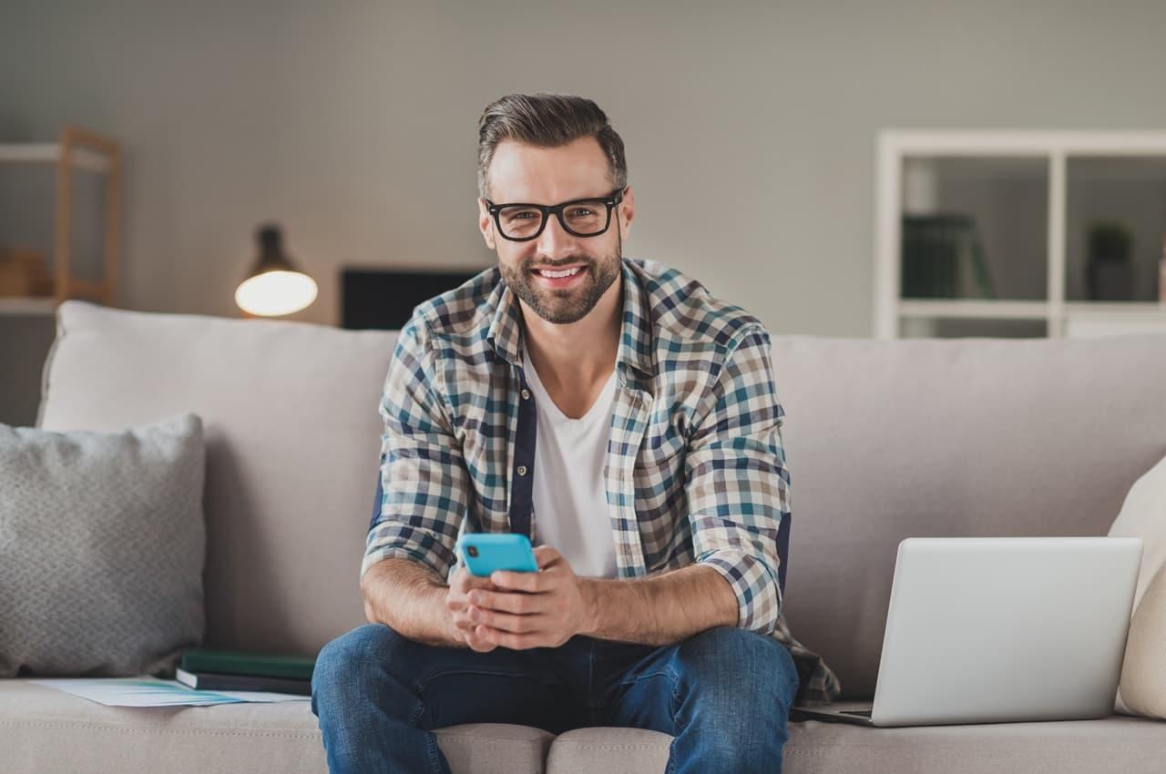 Homem sorridente de barba e óculos sentado no sofá, segurando um celular azul.