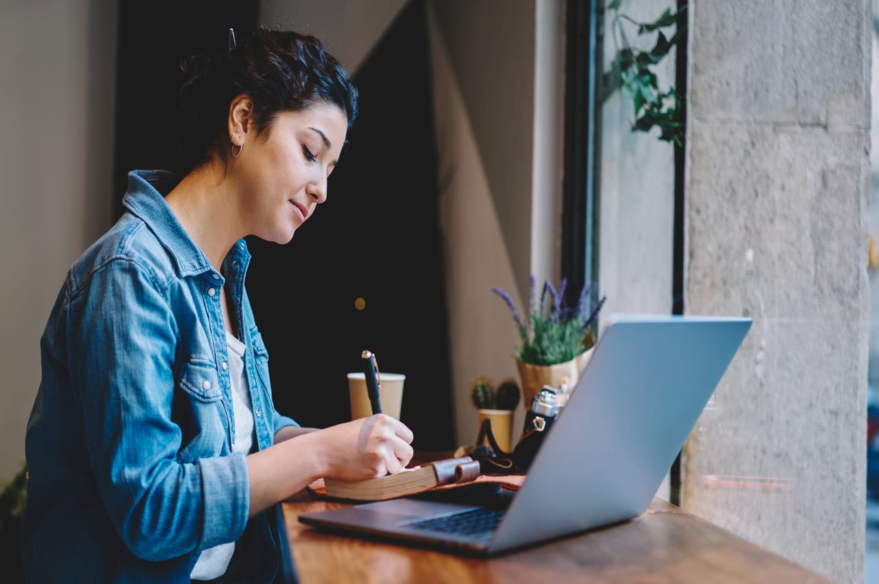 A imagem mostra uma mulher usando seu laptop e fazendo anotações em um bloco de notas.