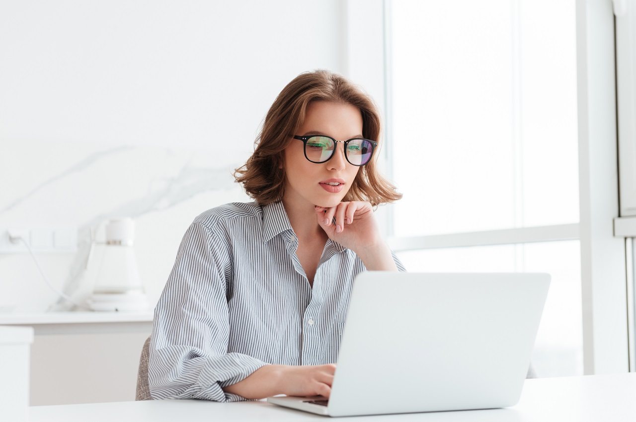 A imagem mostra uma mulher de cabelos curtos e usando uma camisa listrada, sentada em uma mesa, usando seu laptop.