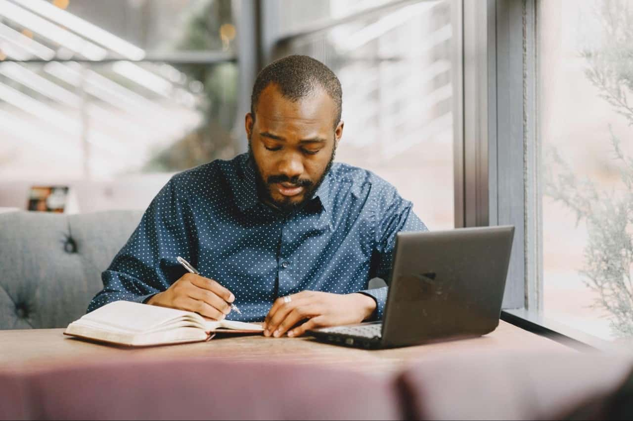 Homem com expressão concentrada anotando em um caderno e com o computador ligado.