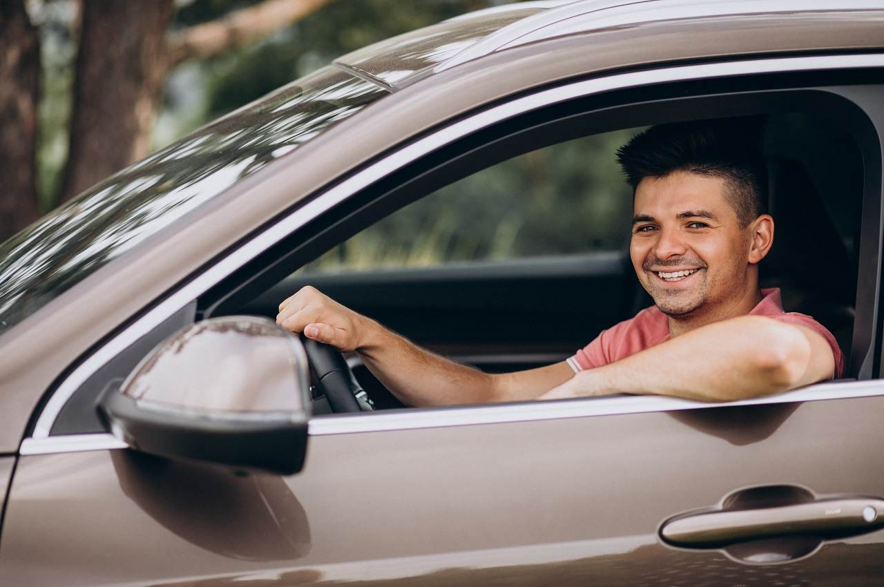 Homem sorridente de camisa vermelha clara, segurando o volante de um carro e o dirigindo.