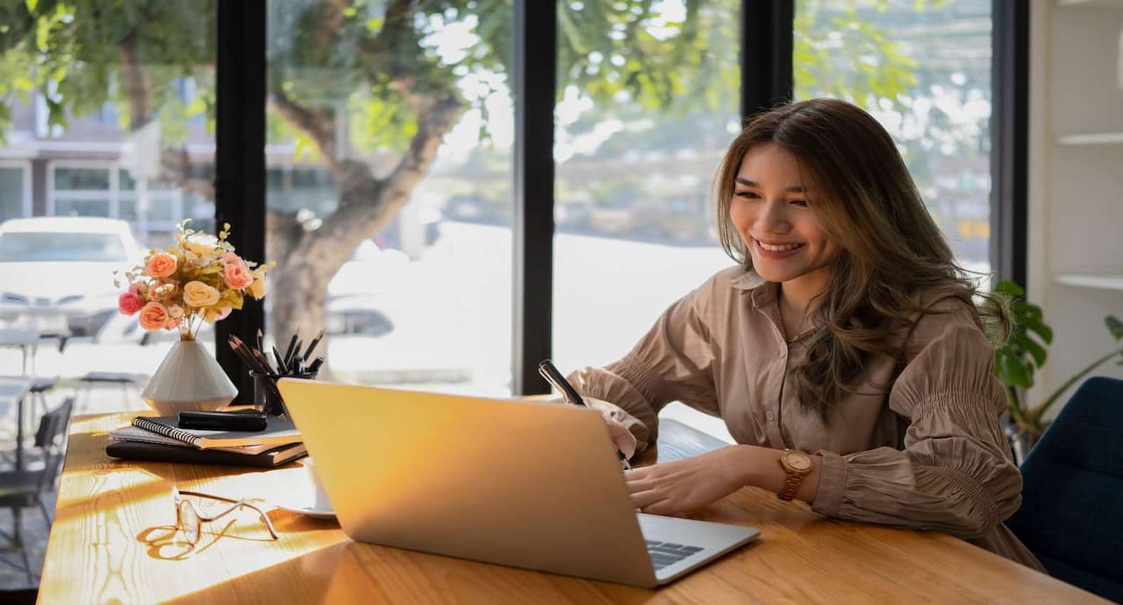 A imagem mostra uma mulher sorridente, usando seu laptop em um escritório. Acima da mesa, um vaso de flores, alguns cadernos e um óculos de grau. 