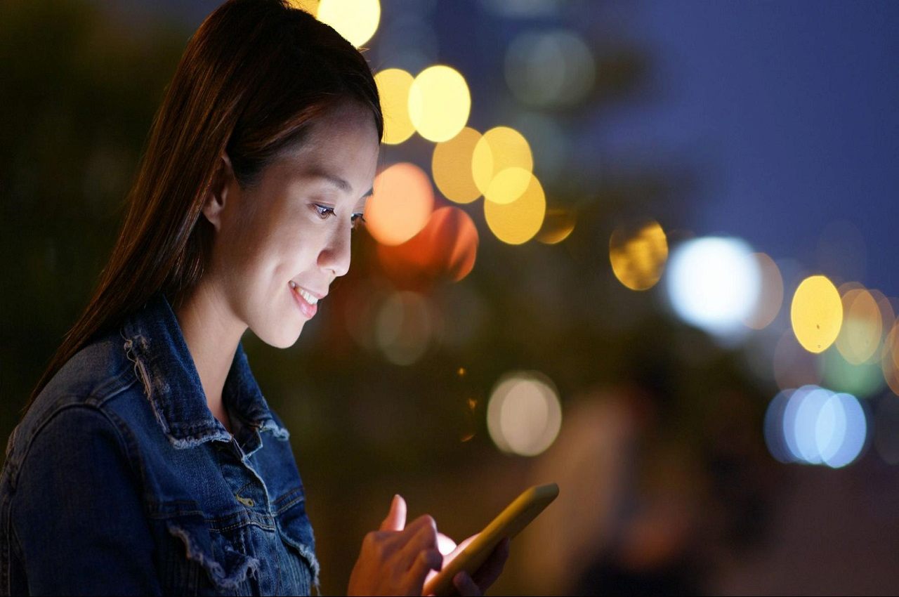 Mulher sorridente utilizando celular e em pé na rua.