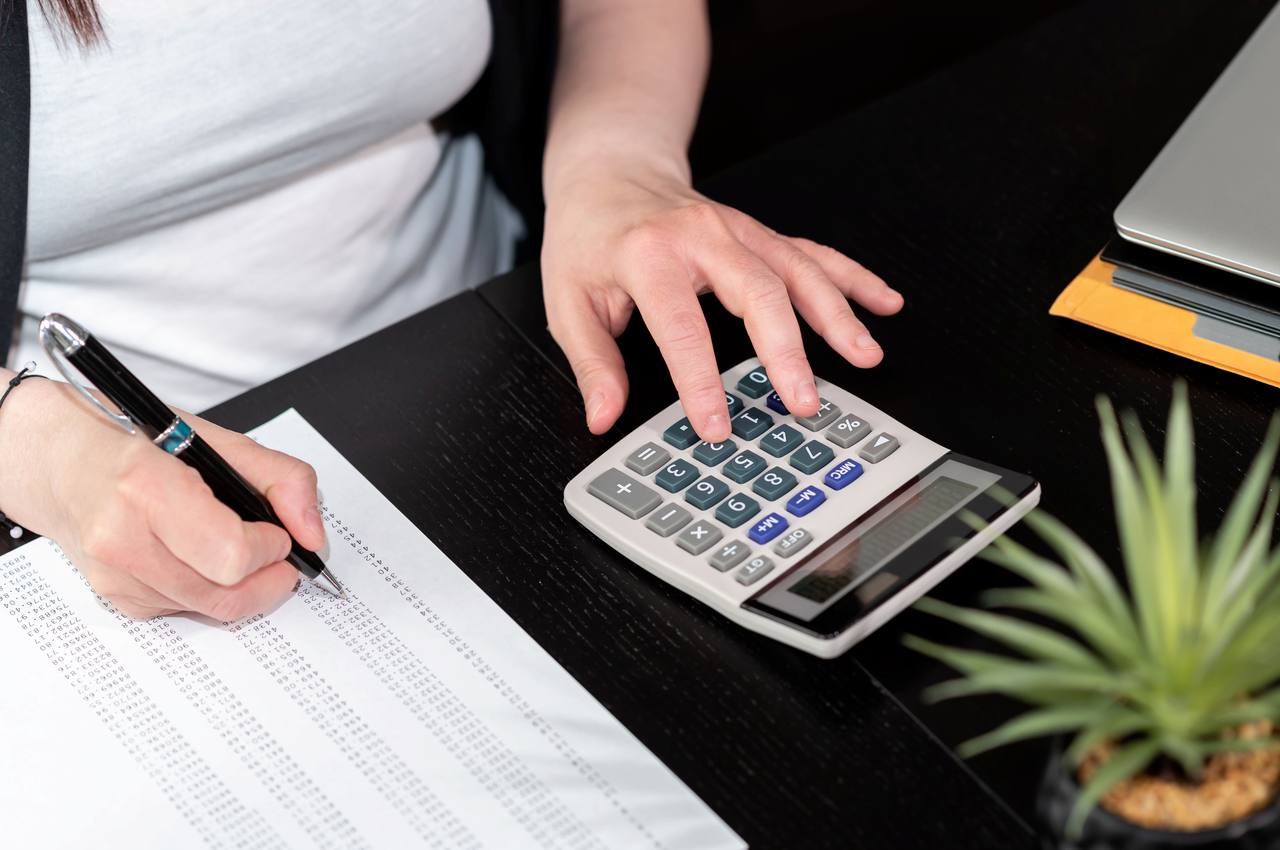 Mulher (seu rosto não aparece na imagem) utilizando calculadora e marcando com uma caneta porcentagens financeiras em uma tabela de papel