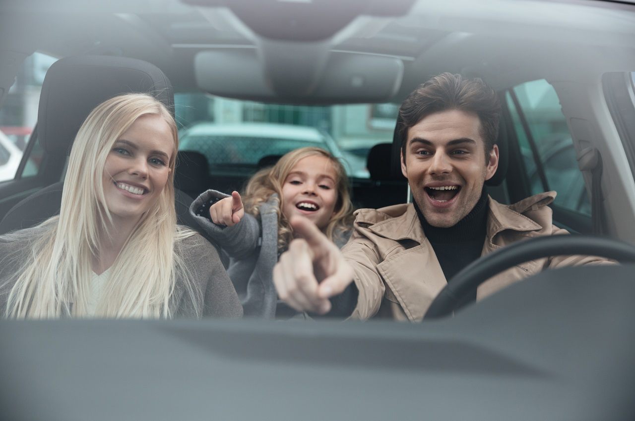 Família sorridente composta por um homem, mulher e uma criança pequena em um veículo, enquanto o pai dirige o carro e aponta para algo à frente.