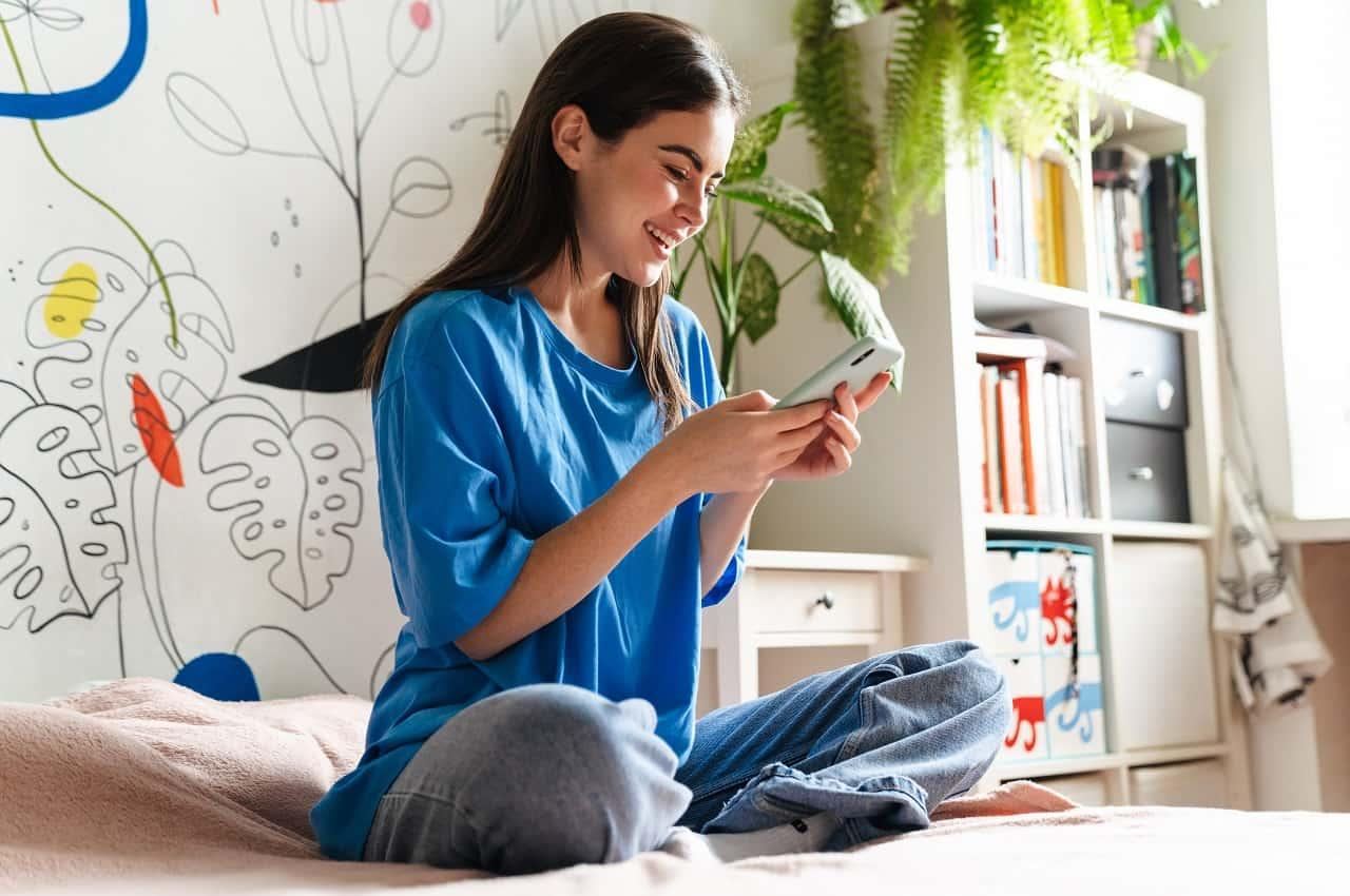 Mulher de cabelos castanhos e sorridentes, usando um pijama de moletom e sentada na cama, enquanto digita no celular.