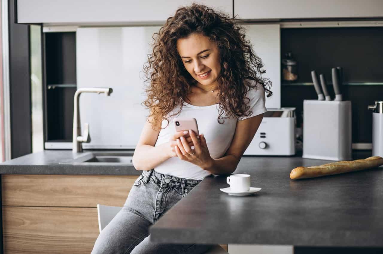 Mulher sorridente de cabelos cacheados longos, apoiada na bancada da cozinha, usando celular para acessar empréstimo rápido e seguro.