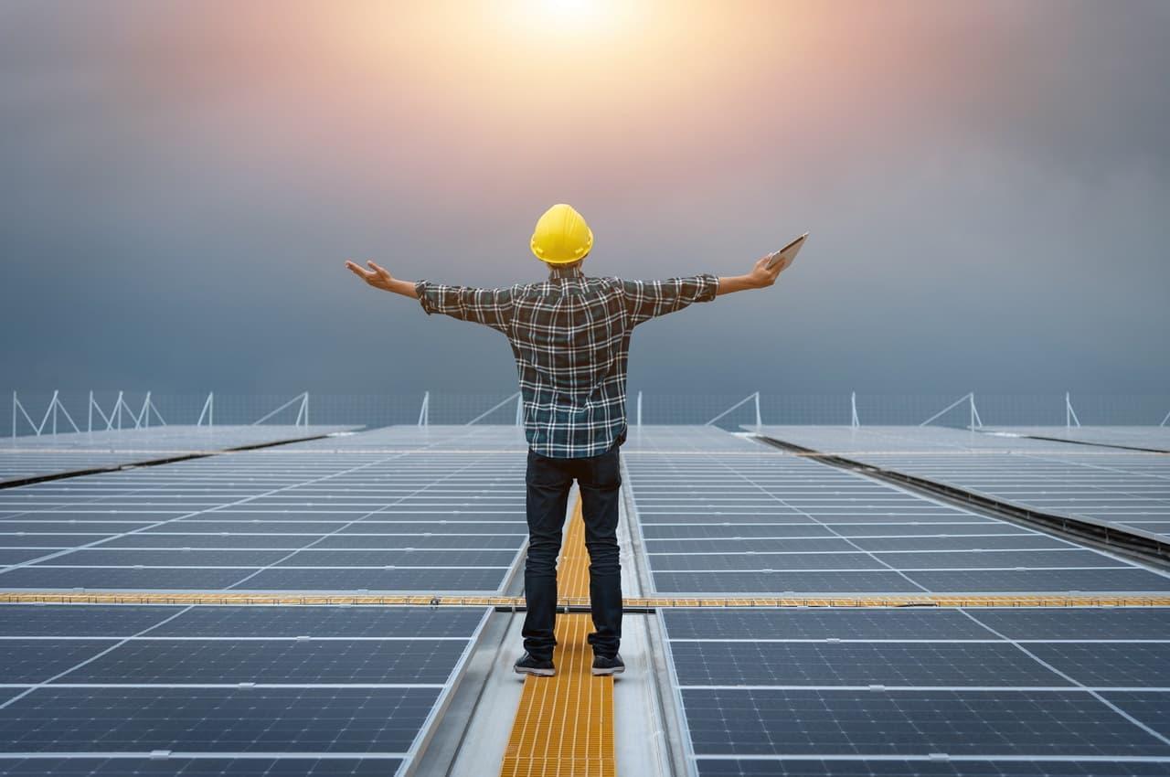 Homem com capacete amarelo de proteção e braços abertos olhando para o sol e em cima de placas solares no chão.