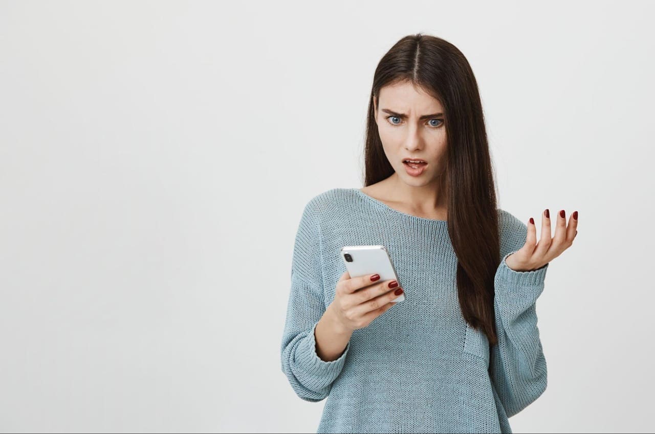 Mulher de longos cabelos lisos castanhos, segura celular com expressão de indignação. A mulher veste um suéter azul-claro e está em pé.