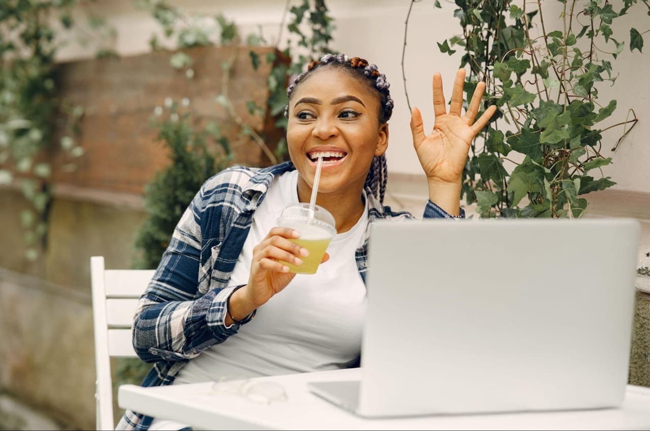 Mulher sorridente bebendo um milkshake e com a mão levantada em cumprimento. 