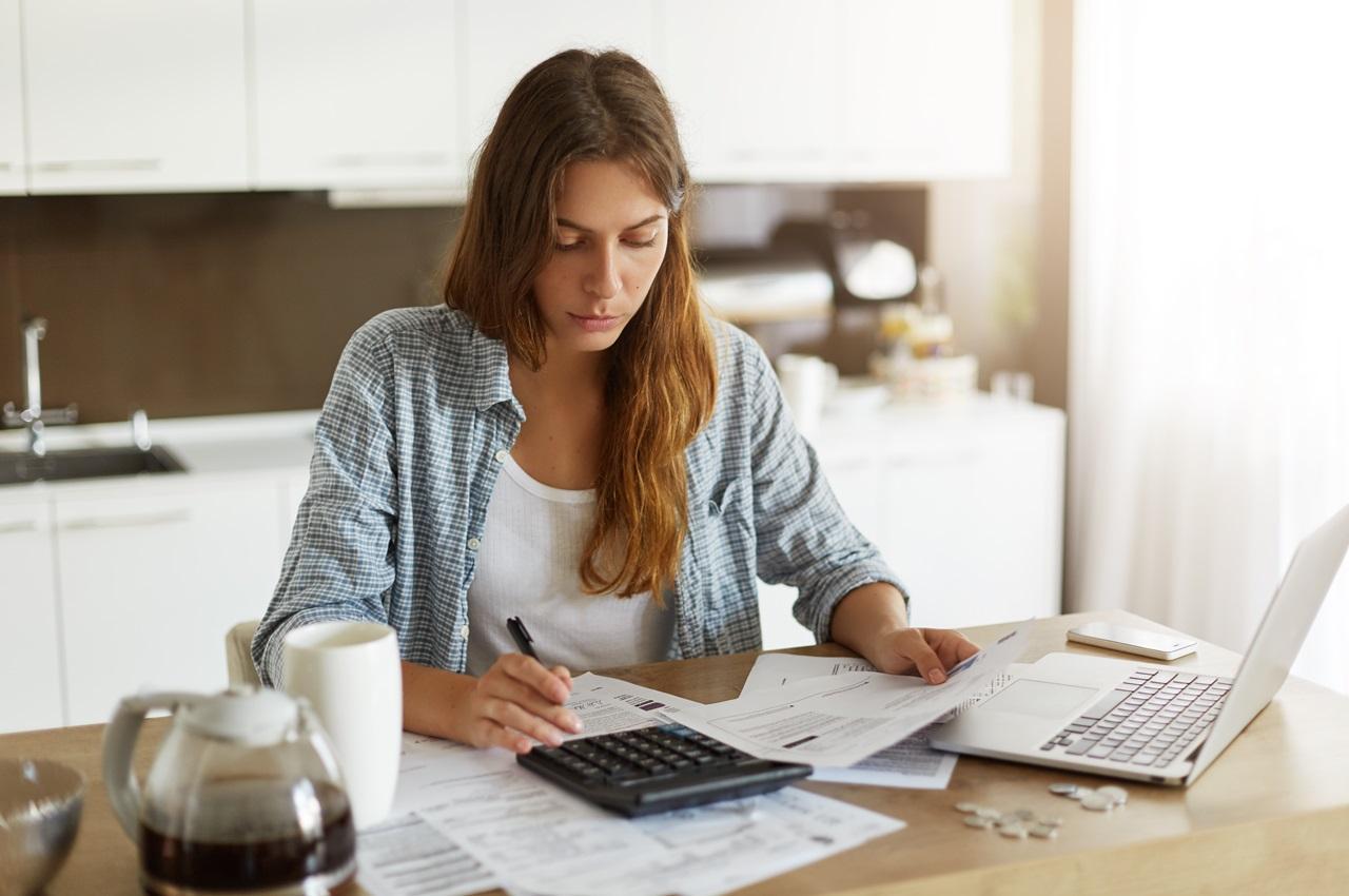 Mulher de longos cabelos castanhos claros e expressão concentrada, segurando papéis de contas e uma calculadora.