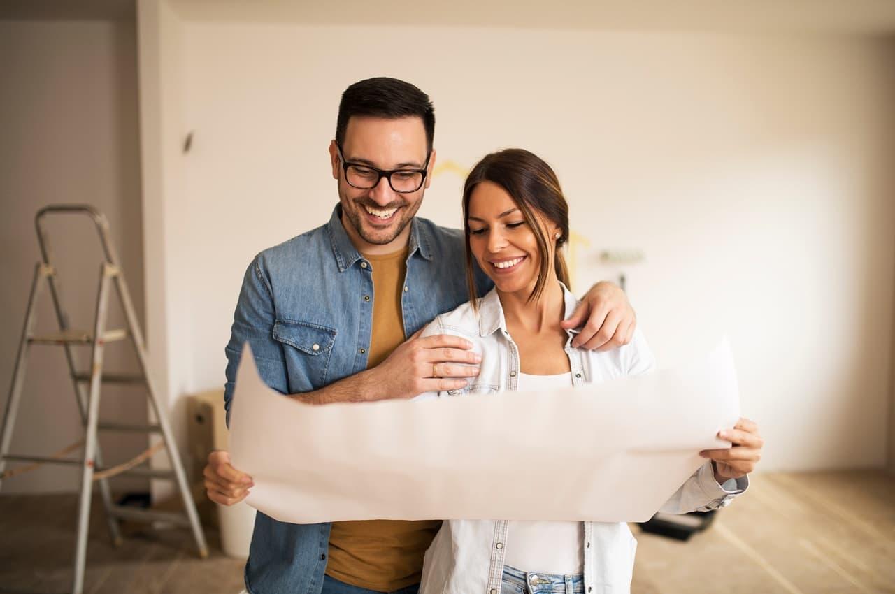 Casal de homem e mulher sorridentes olha a planta de reforma da casa e, ao fundo, tem materiais de construção.